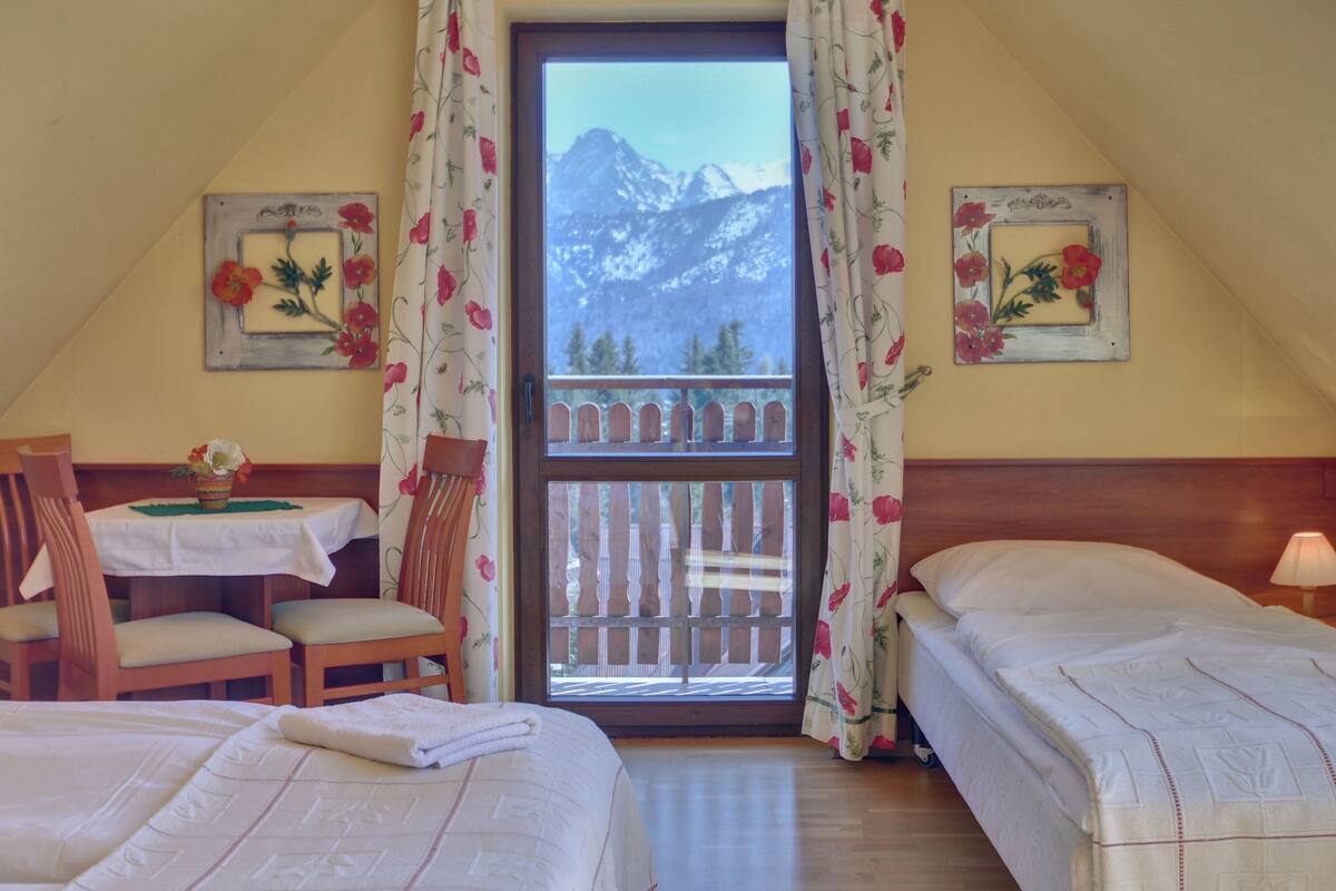 Una habitación con dos camas, una mesa de madera con un florero, y vistas a las montañas a través de una ventana.