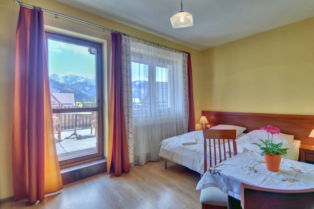 Habitación de hotel con dos camas, un cabecero de madera, una mesita con una planta y un balcón con vista a las montañas.
