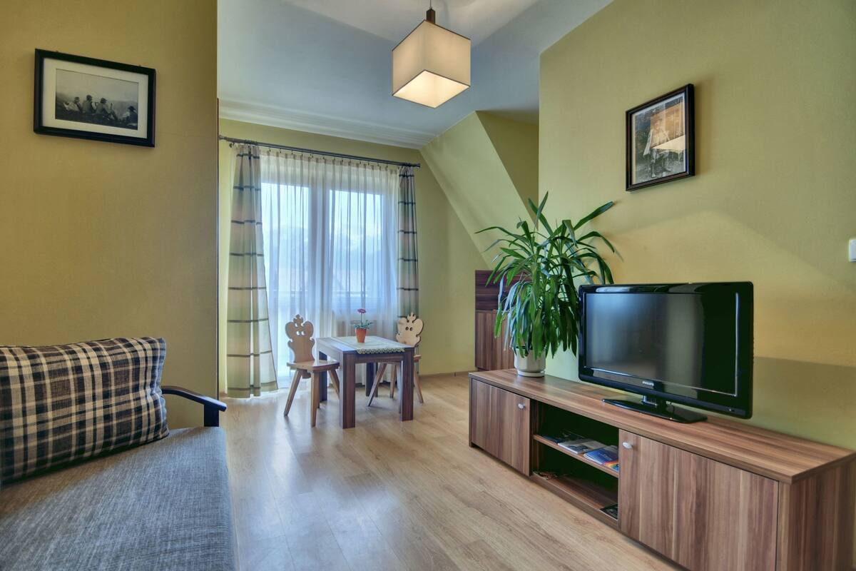 Una sala de estar con sofá, televisor sobre un mueble de madera, mesa de comedor con sillas y una planta grande en maceta.