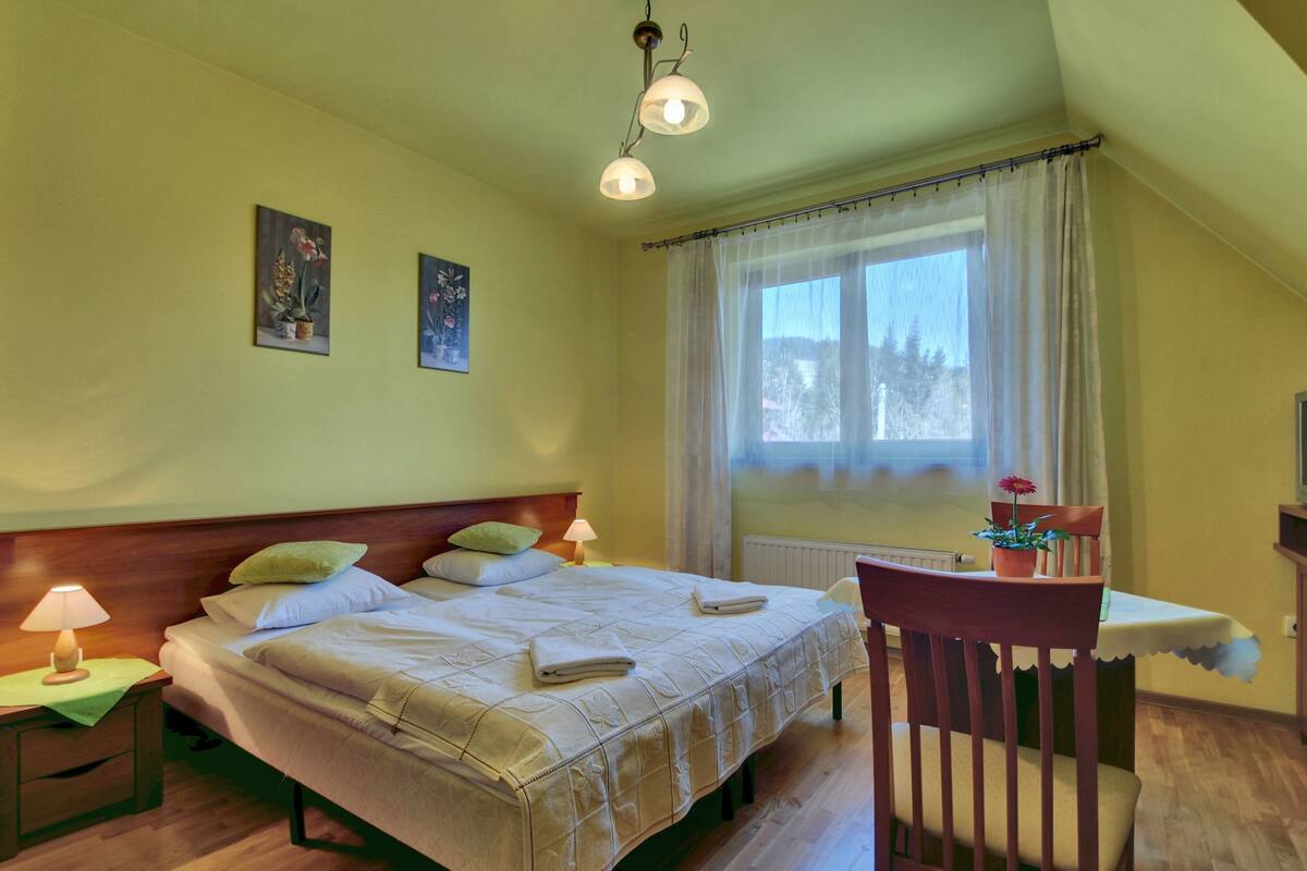 Un dormitorio con una cama, dos mesitas de noche, una silla, un televisor y una ventana con cortinas.