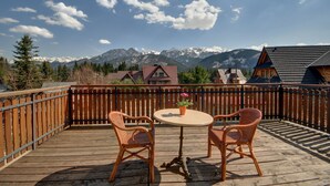 Una terraza de madera con dos sillas de mimbre y una mesa redonda, con vistas a una cordillera de montañas con picos nevados y casas al fondo.