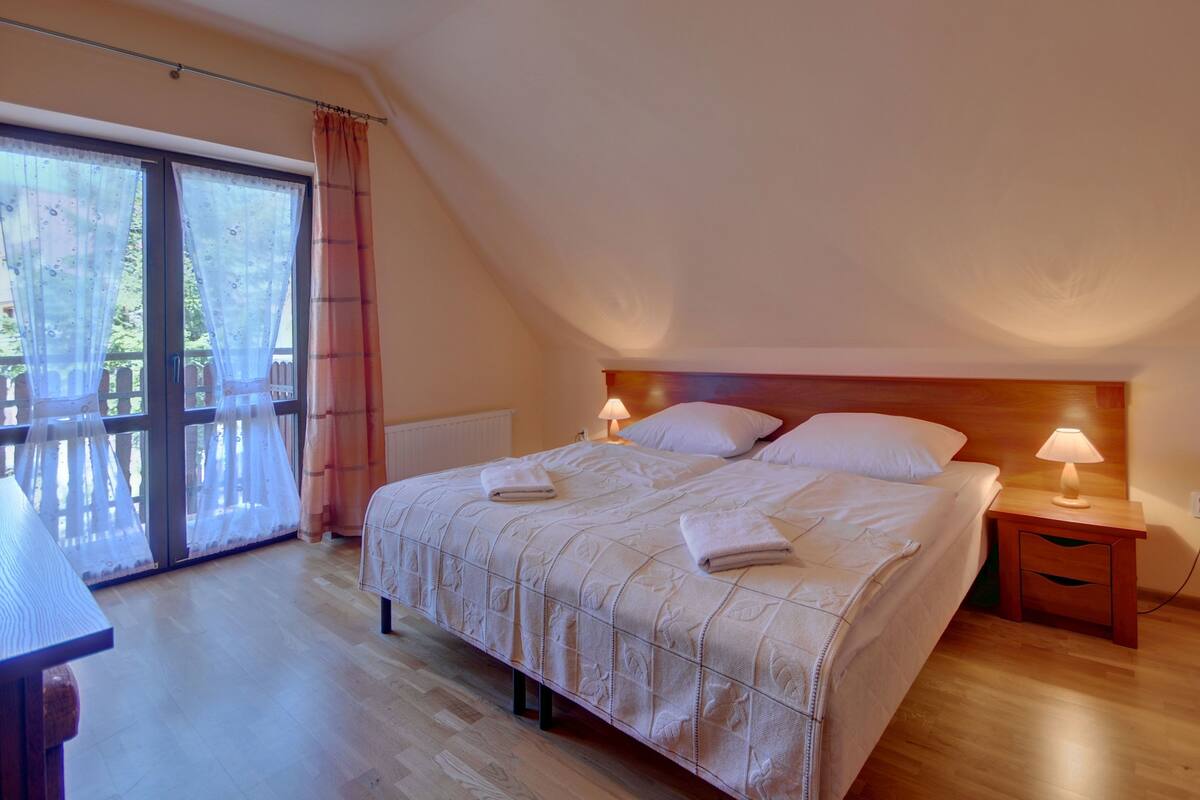 Un dormitorio con una cama grande, dos mesitas de noche con lámparas, un cabecero de madera y vistas a un balcón a través de una puerta de cristal.