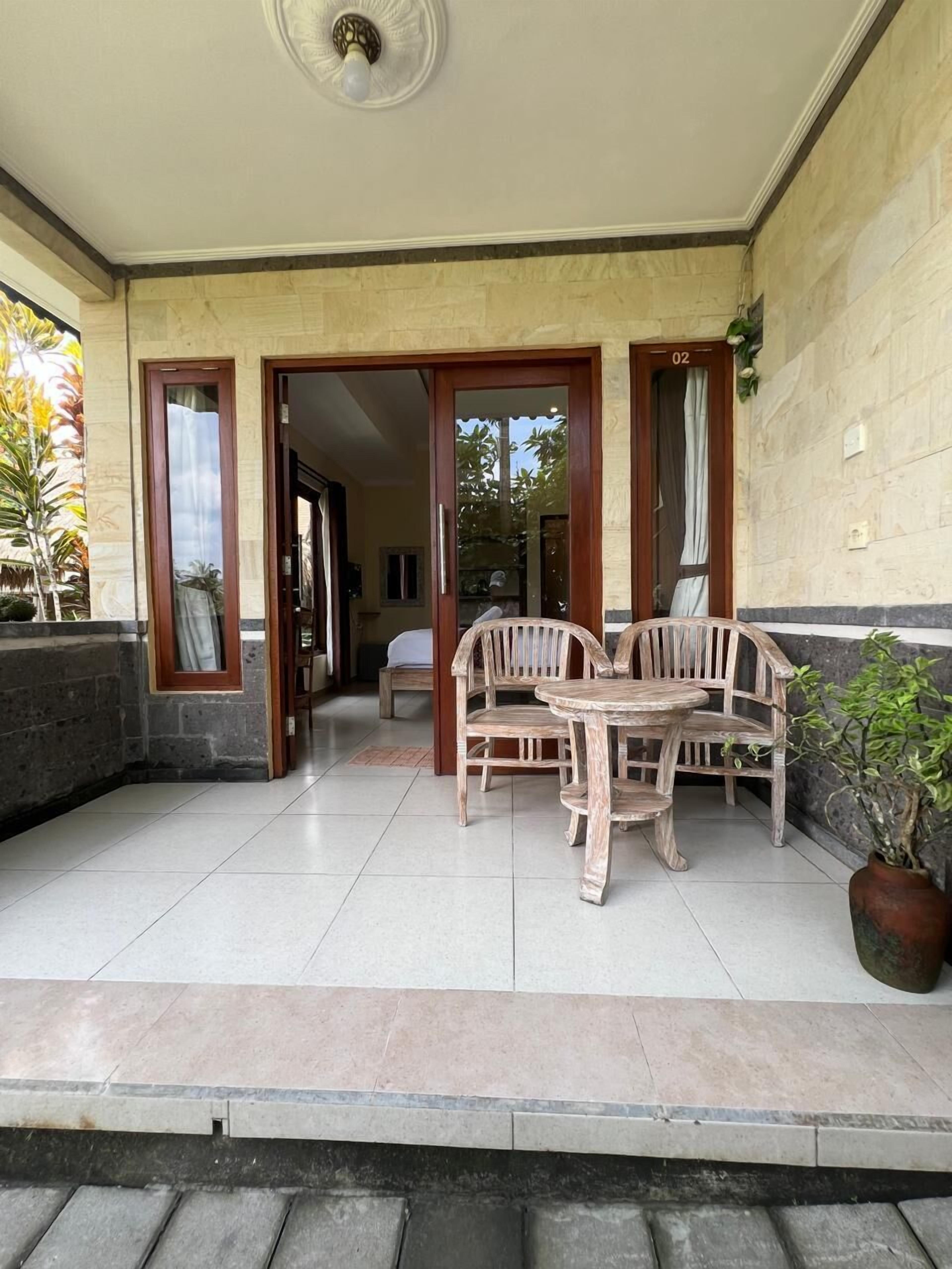 A covered outdoor seating area with a wooden table and chairs, a potted plant, and a door leading inside.