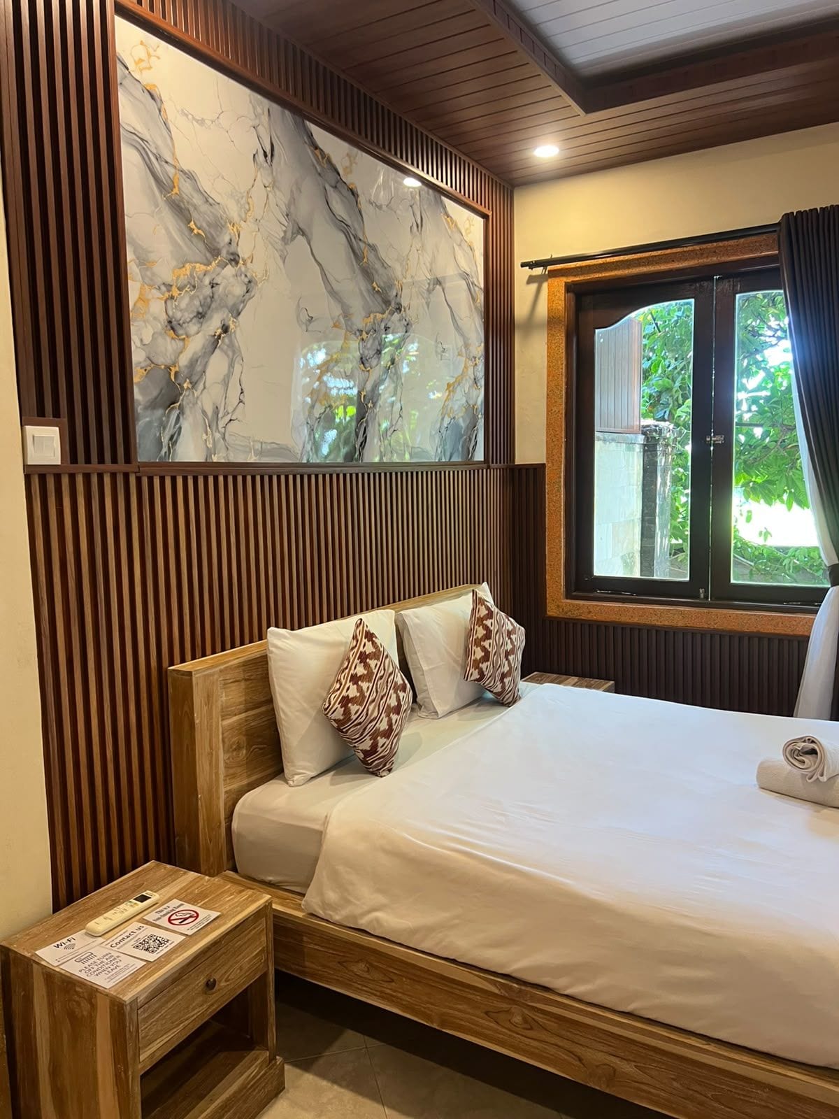 Une chambre d’hôtel comprenant un lit, une tête de lit en bois, une œuvre d’art en marbre, une fenêtre avec des rideaux et une petite table de chevet sur laquelle se trouve un appareil numérique.