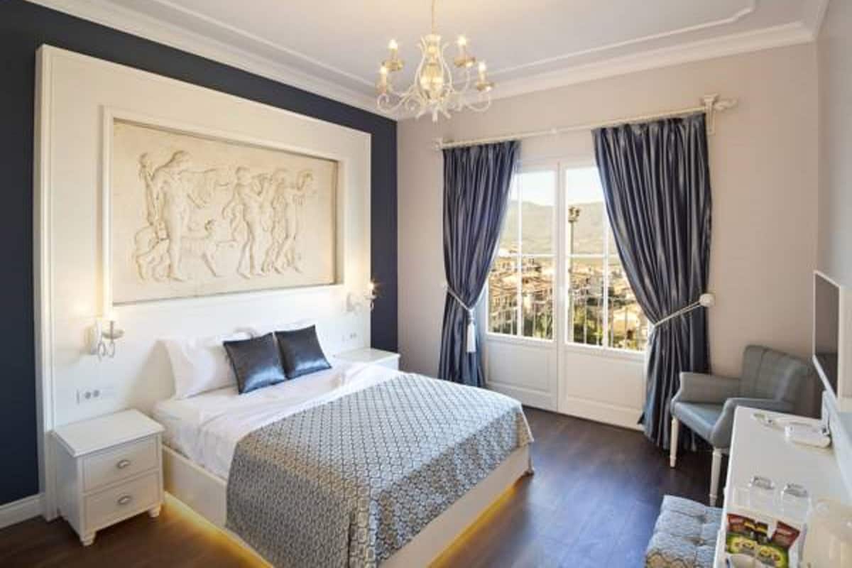 Un dormitorio con una cama grande, una mesita de noche, un escritorio, una silla, una araña y una ventana con cortinas.