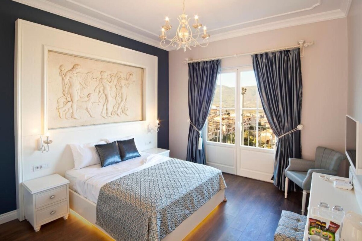 A bedroom with a large bed, a nightstand, a desk, and a view of the city through the window.