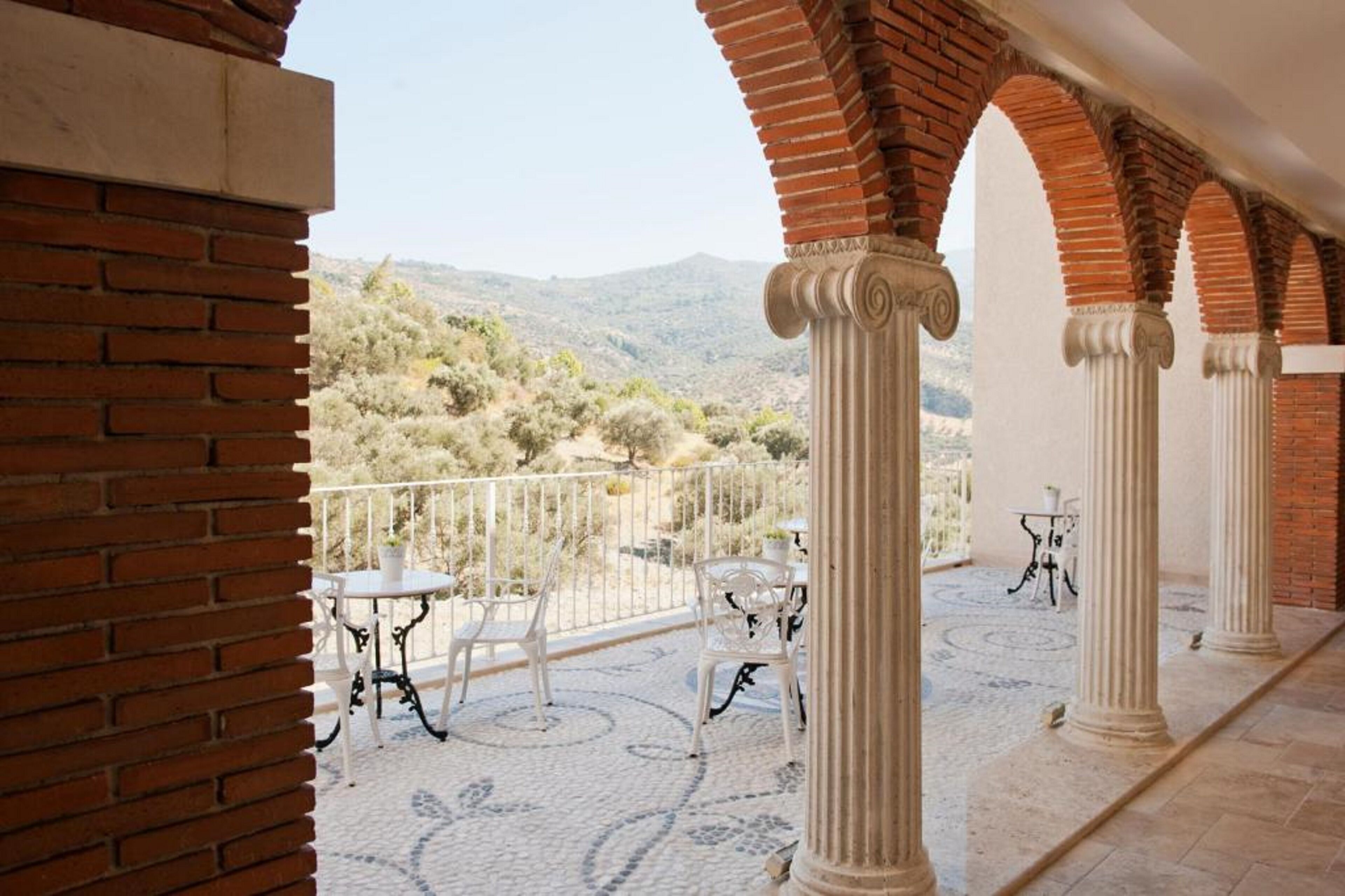 A covered outdoor area with brick columns, white chairs, and tables, overlooking a hilly landscape.