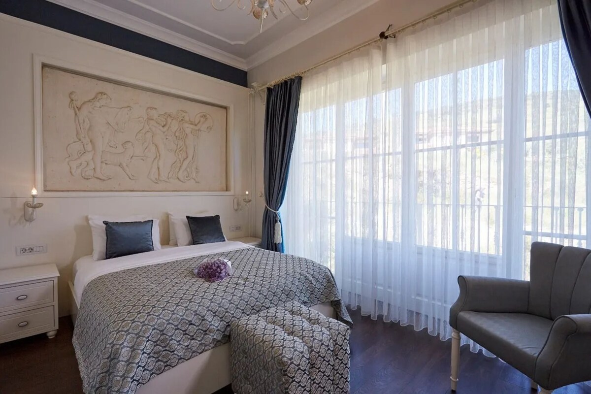 A bedroom with a bed, a chair, a nightstand, a wall-mounted artwork, and a large window with sheer curtains.