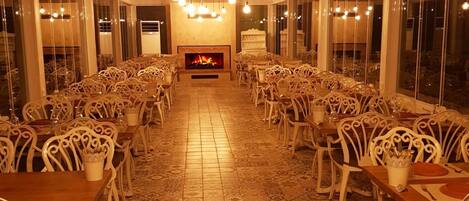 Un comedor amplio con sillas y mesas blancas de detalles ornamentales, suelo estampado y chimenea en el centro.