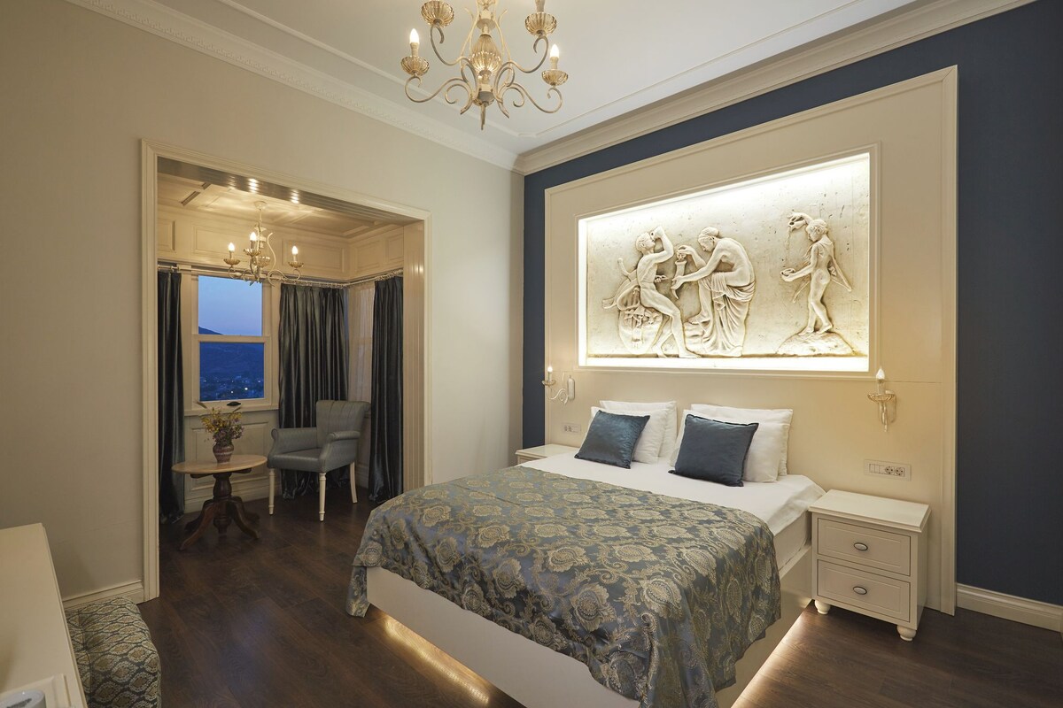 A bedroom with a bed, bedside tables, a chandelier, a wall-mounted sculpture, and a view of the outdoors.