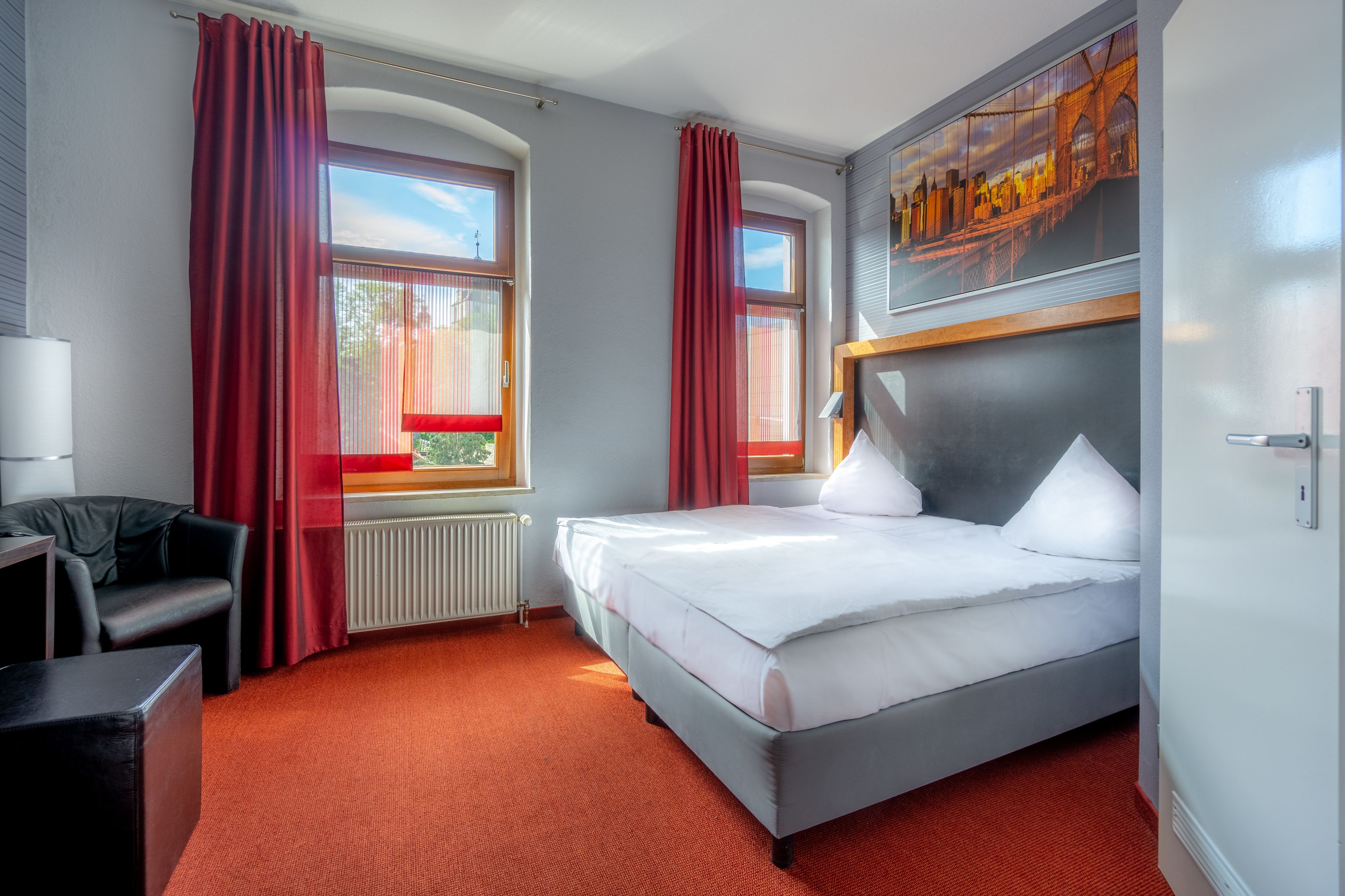 A hotel room with a bed, a desk, a chair, and a window with red curtains.