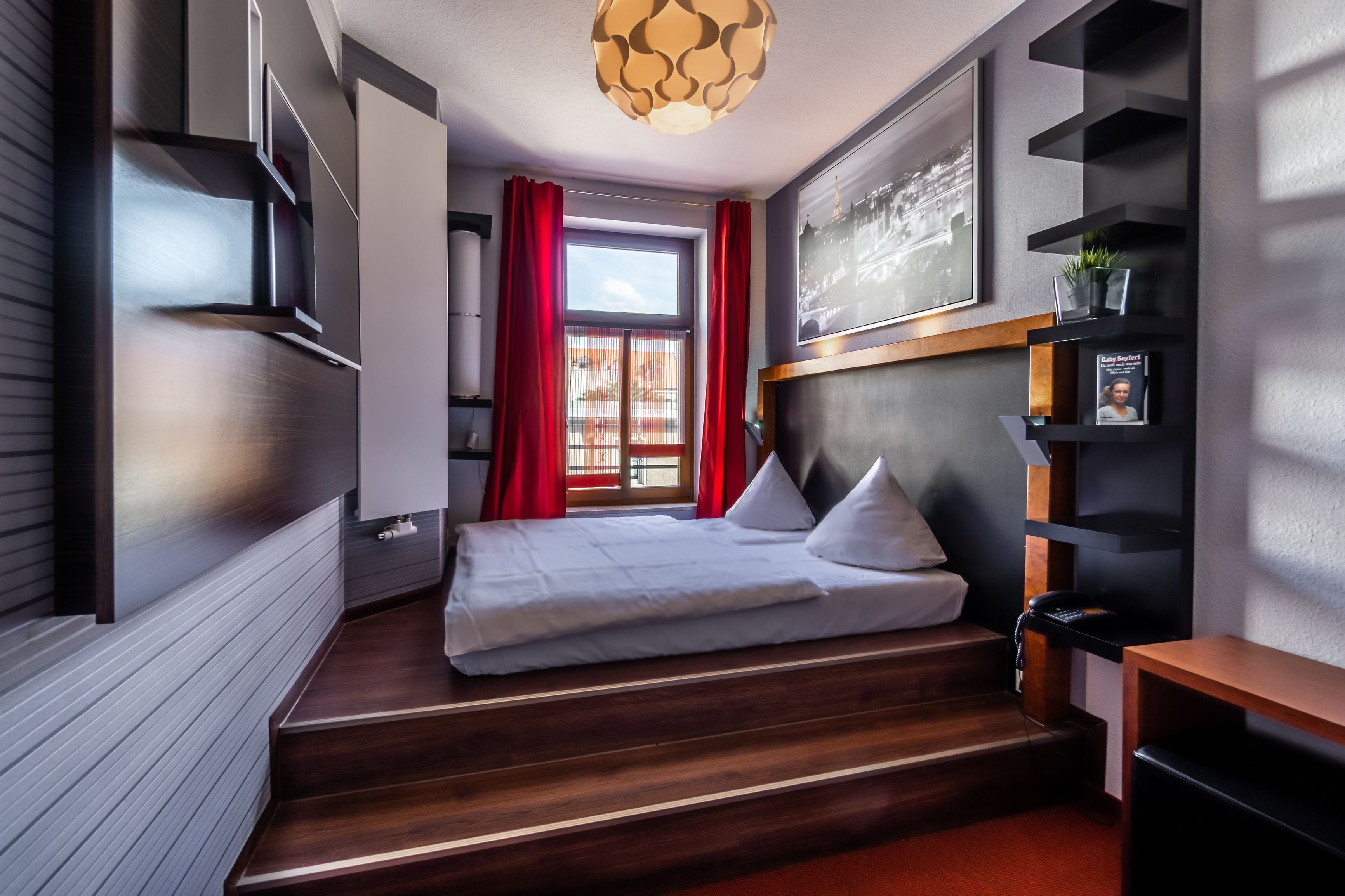A modern bedroom with a bed, red curtains, a wooden staircase, a desk, and a wall-mounted shelf.