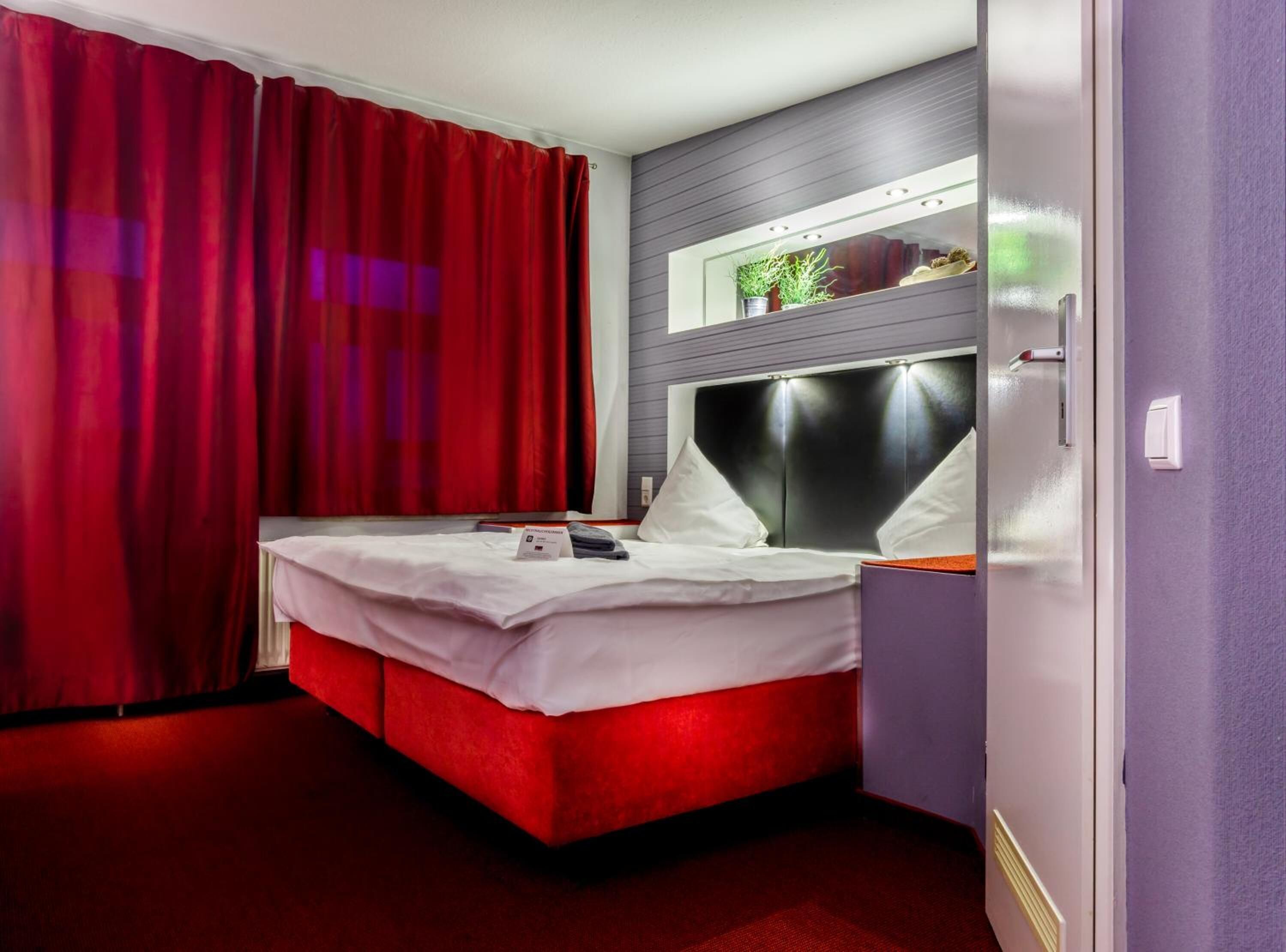 Ein Hotelzimmer mit einem roten Bett, weißer Bettwäsche, einem modernen Kopfteil und einem kleinen Fenster mit Pflanzen.