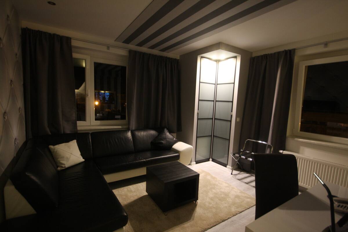 A modern living room with a black leather sofa, a small black ottoman, a white armchair, and a glass partition wall.