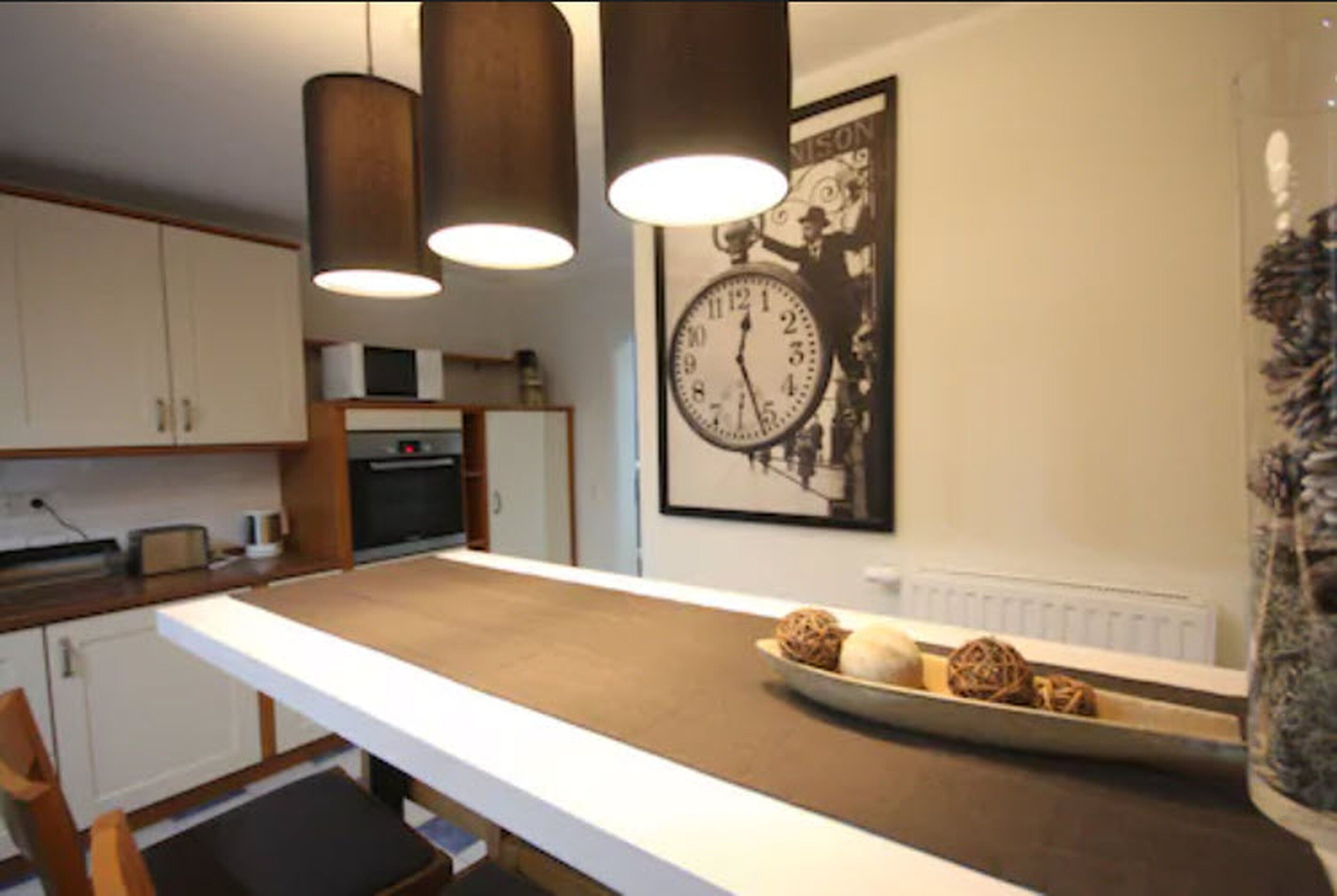 A kitchen with a long counter, hanging pendant lights, a clock, and decorative items on the counter.