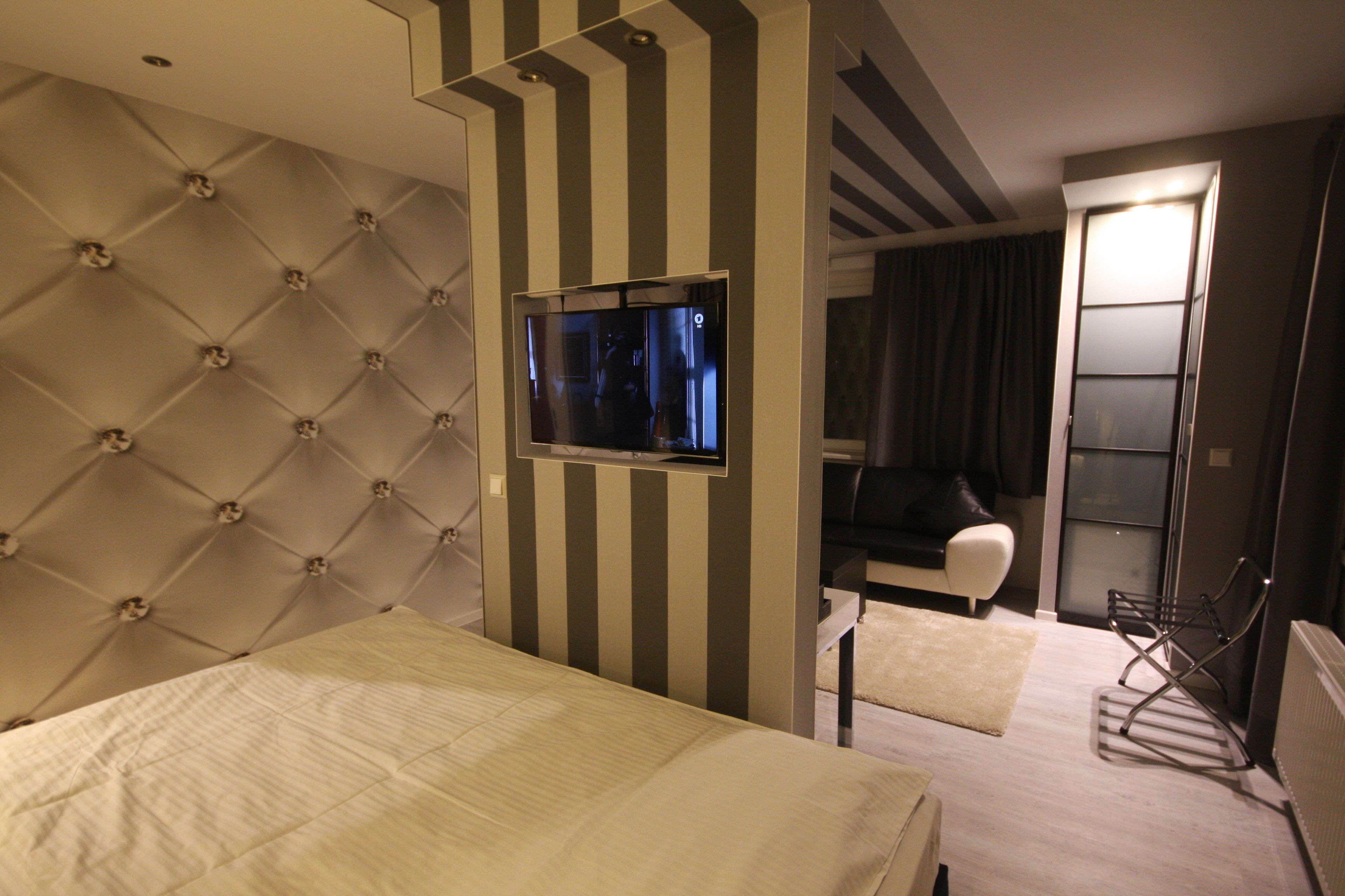 Habitación de hotel con cabecera capitonada, televisor de pantalla plana montado en una pared a rayas, sofá, sillón y radiador.