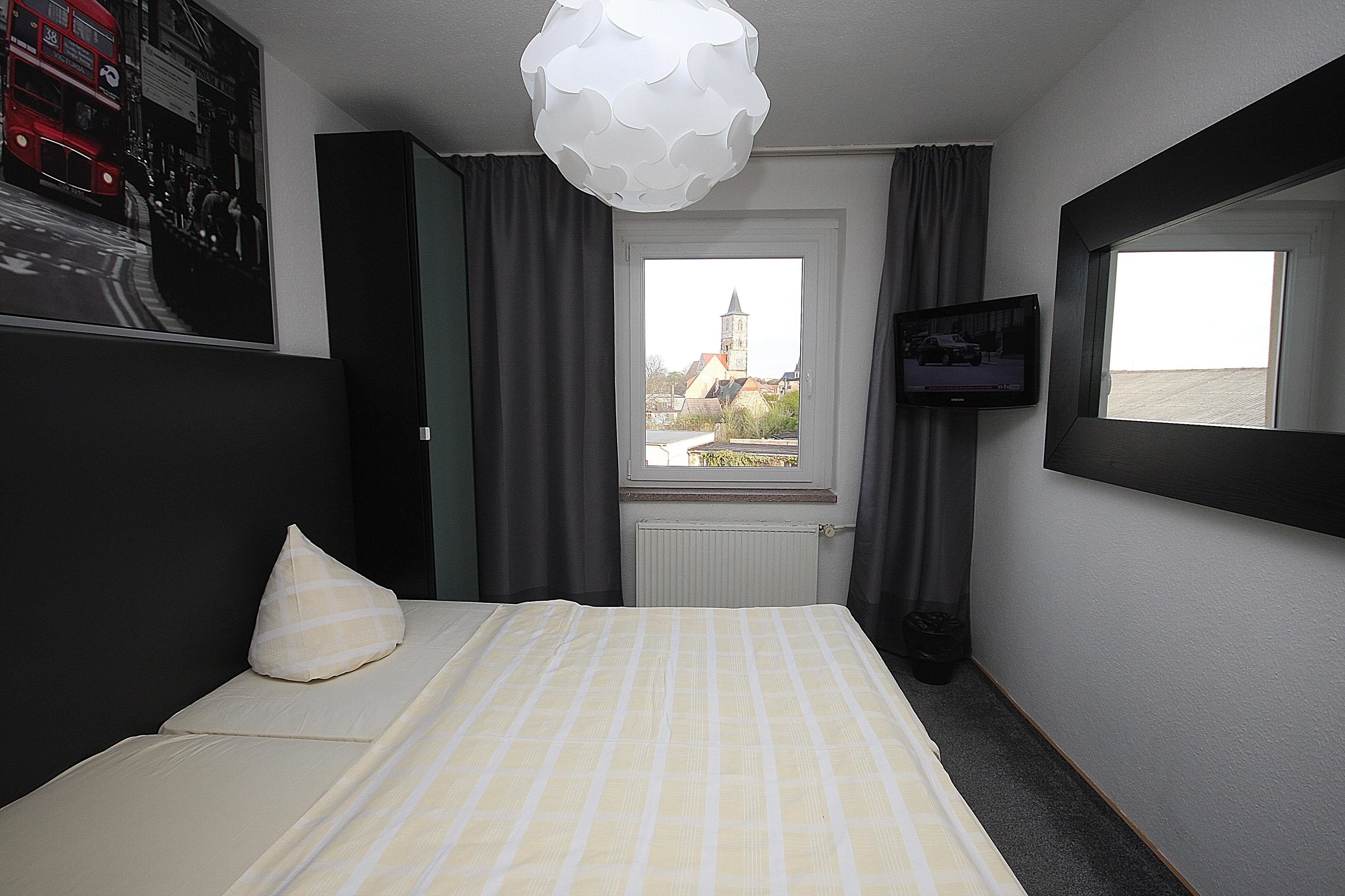 A hotel room with a bed, a television, a window with a view of a building, and a decorative ceiling light.