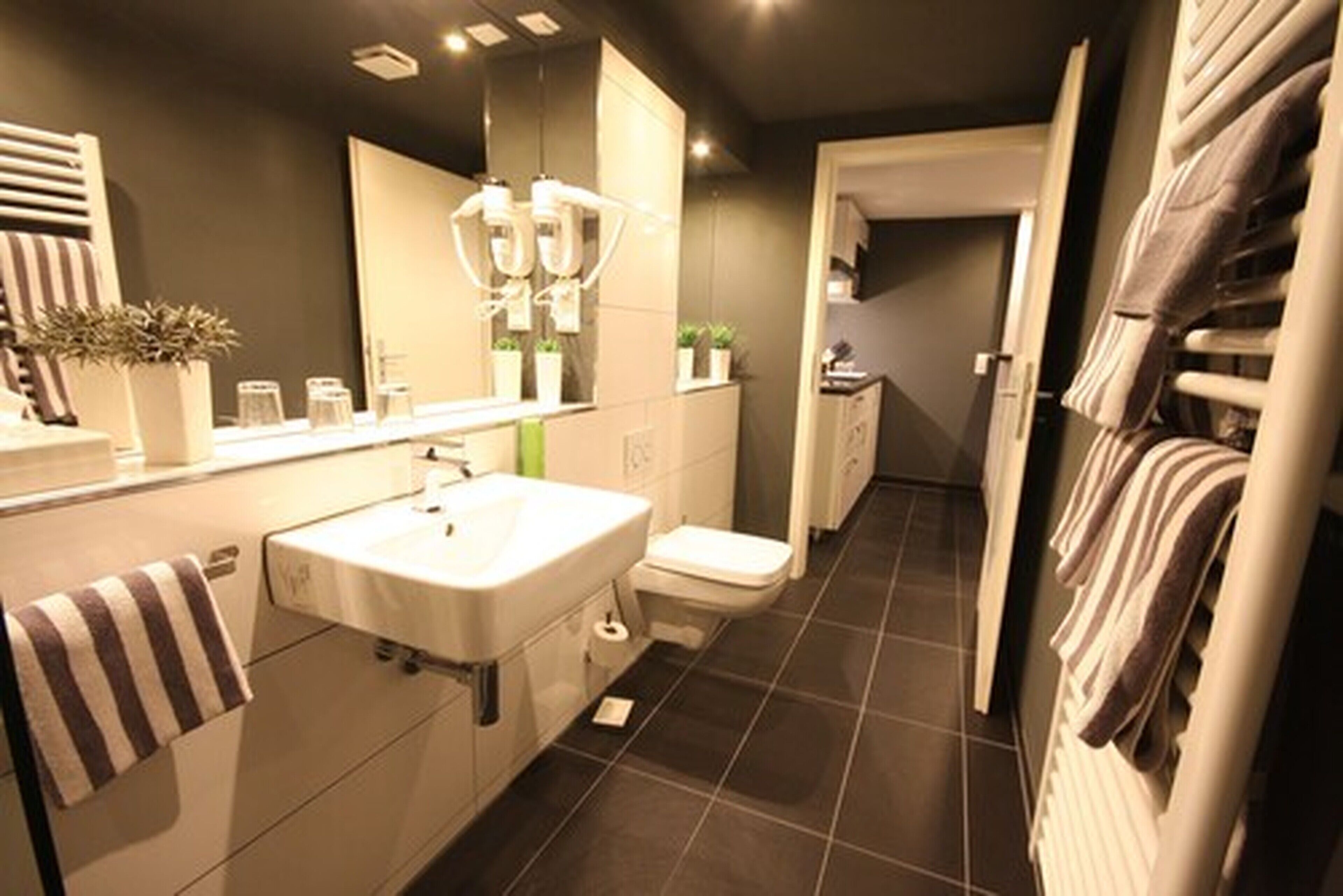 A modern bathroom with a large mirror, a sink, and a toilet. There are towels hanging on the wall and a plant on the counter.