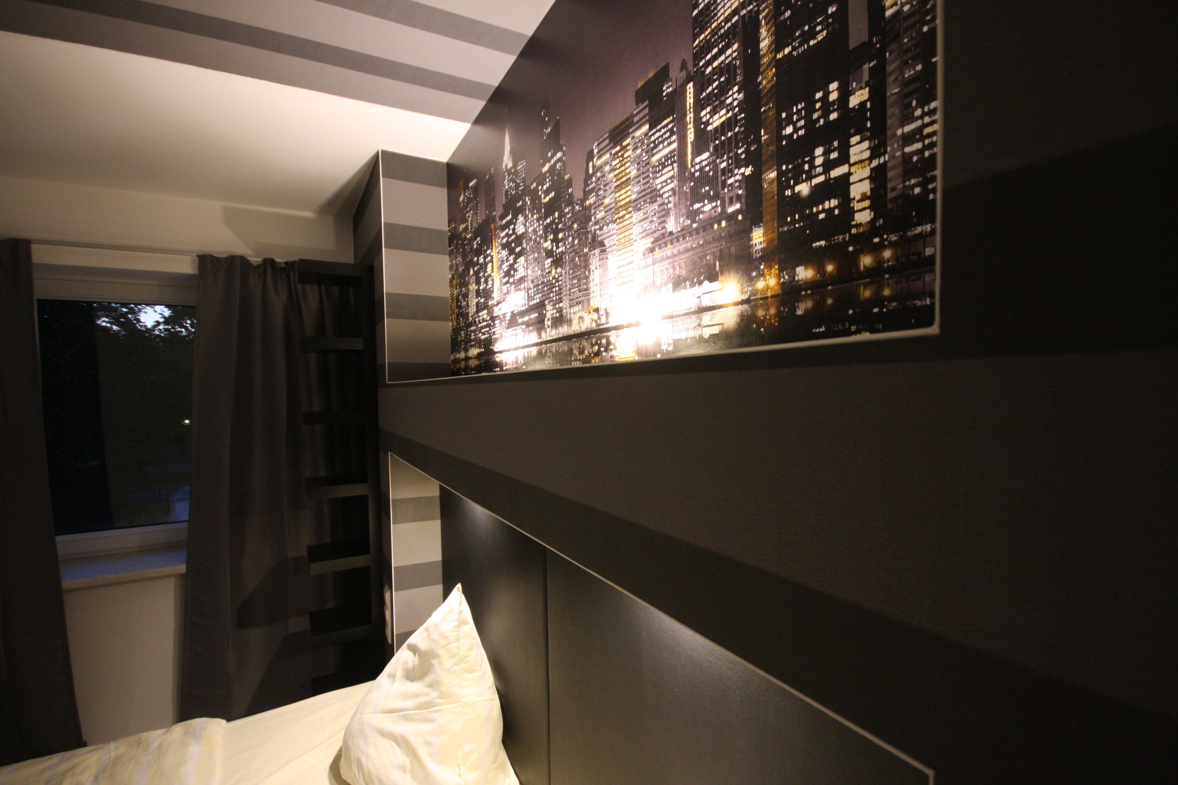 A bedroom with a bed, a nightstand, a wall-mounted shelf, and a cityscape artwork.