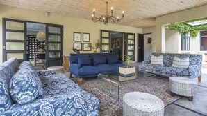 Un vaste espace de vie comprenant un canapé bleu, un canapé à motifs, une table basse en verre et des poufs en osier. On y trouve également un lustre, des œuvres d’art encadrées accrochées aux murs et une porte donnant sur une autre pièce.