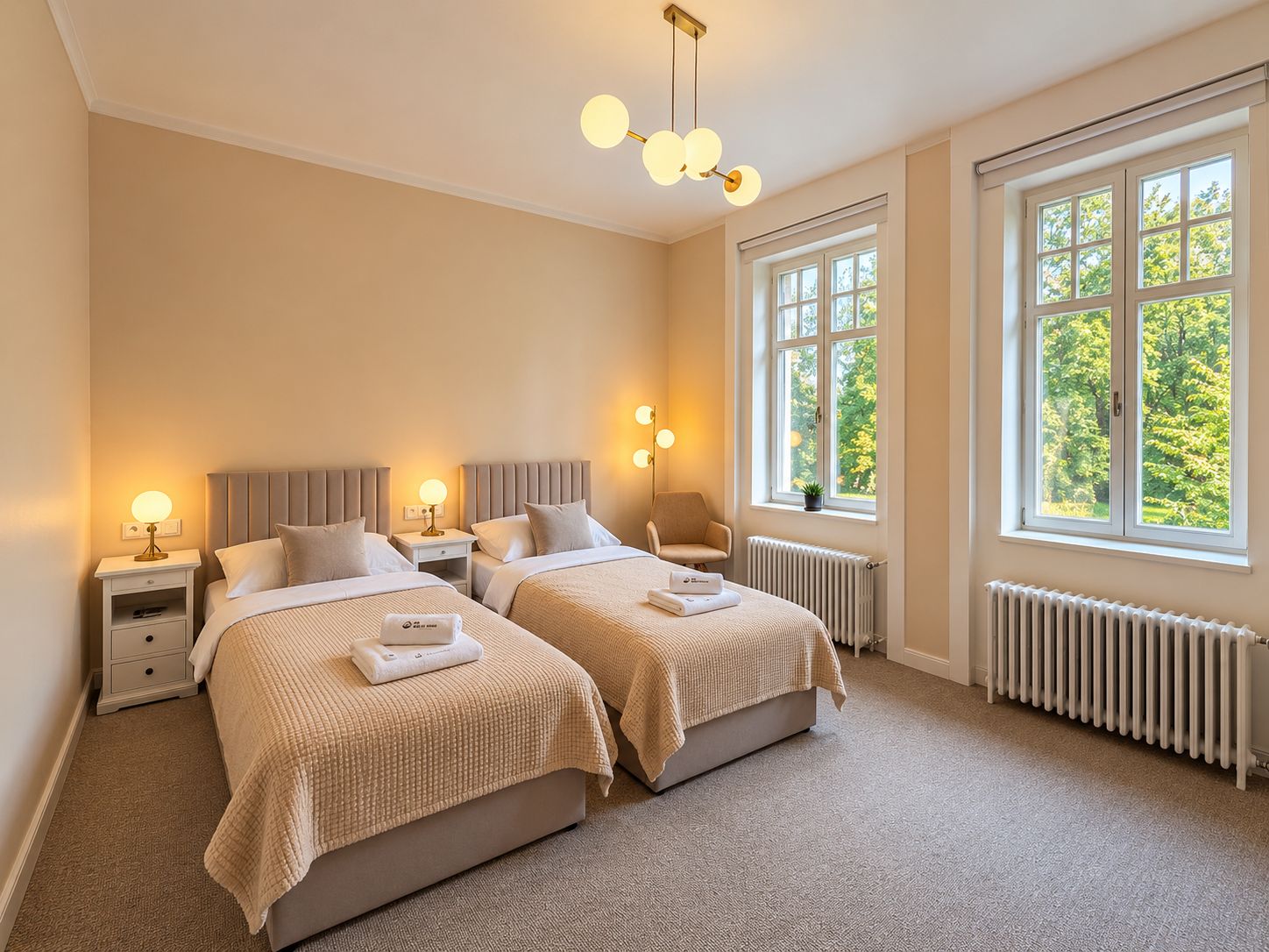 A hotel room with two beds, bedside tables, lamps, a radiator, and large windows with a view of trees.