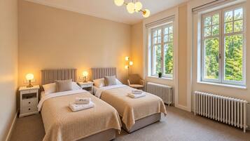 A hotel room with two beds, bedside tables, lamps, a radiator, and large windows with a view of trees.