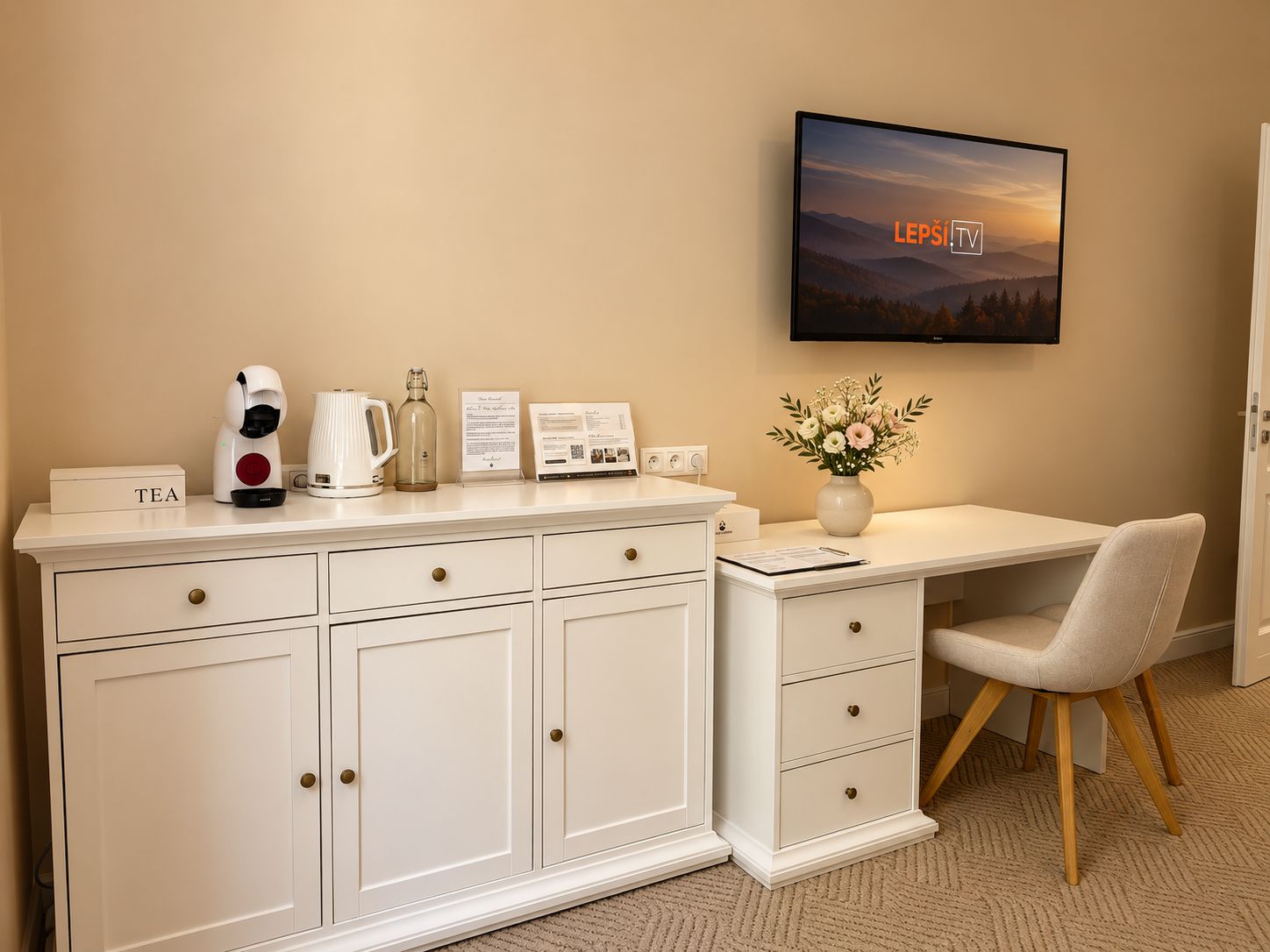 Habitación de hotel con un escritorio blanco, televisor montado en la pared, aparador con cajones, espacio para té y un jarrón con flores.