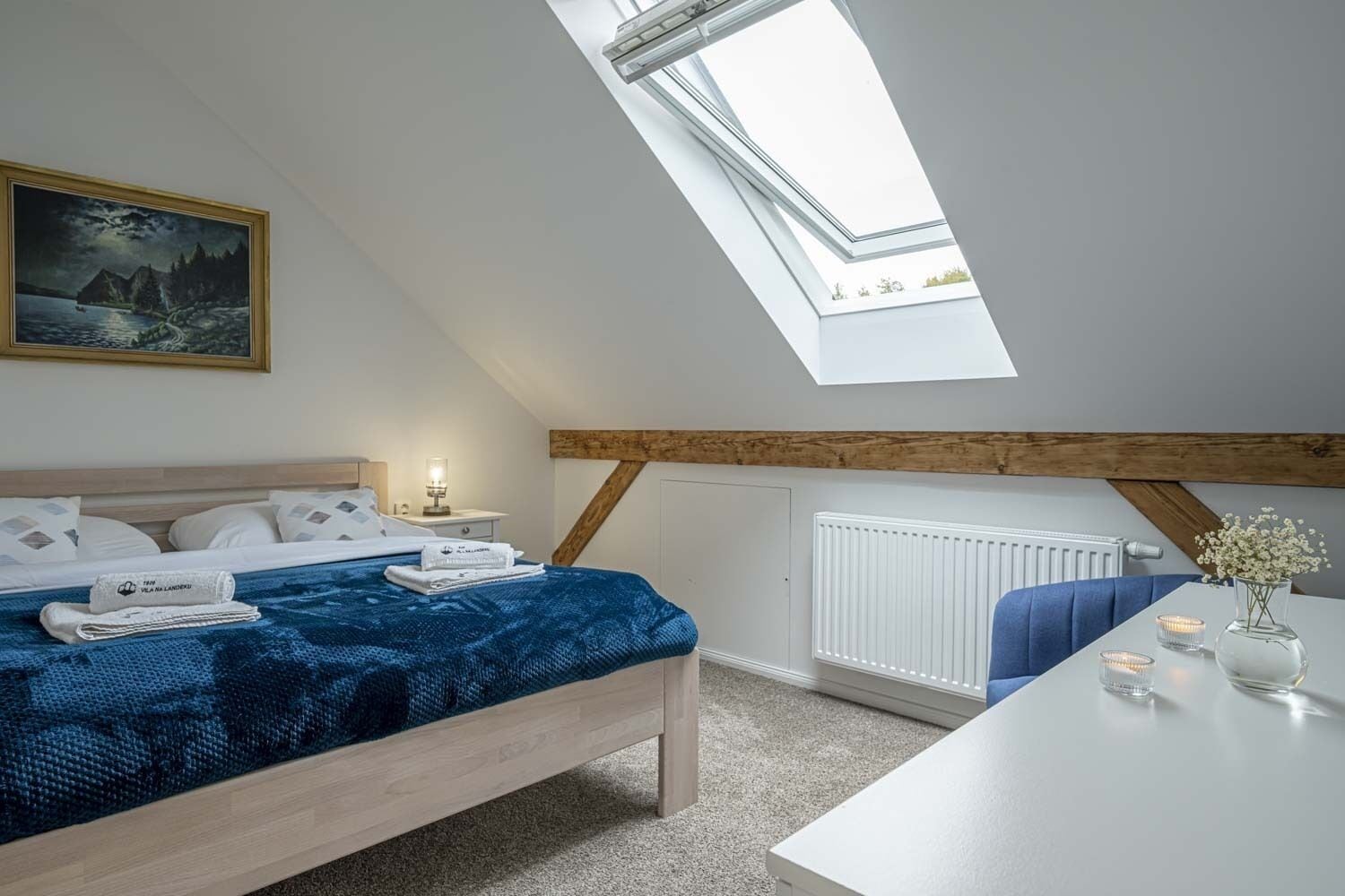Ein Schlafzimmer mit Oberlicht, einem Bett mit blauer Decke, einem Nachttisch mit Lampe und einem Gemälde an der Wand.