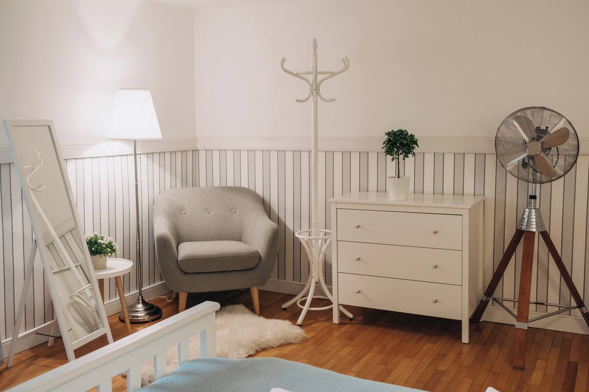 Une chambre à coucher comprenant un lit, une commode, une chaise, une lampe, un porte-manteau, un ventilateur et une petite plante.