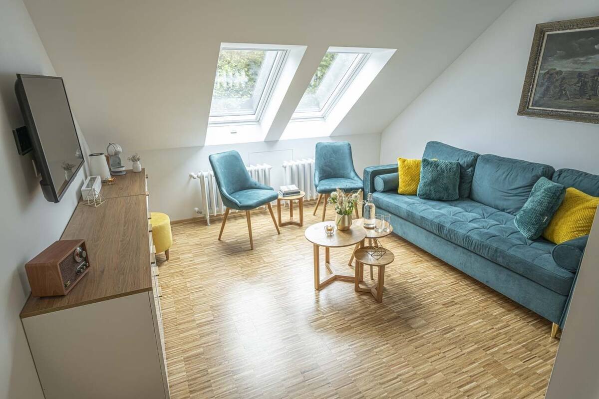 Un appartement moderne de style « loft », avec un canapé bleu, deux fauteuils, une table basse en bois et un téléviseur posé sur un meuble en bois.