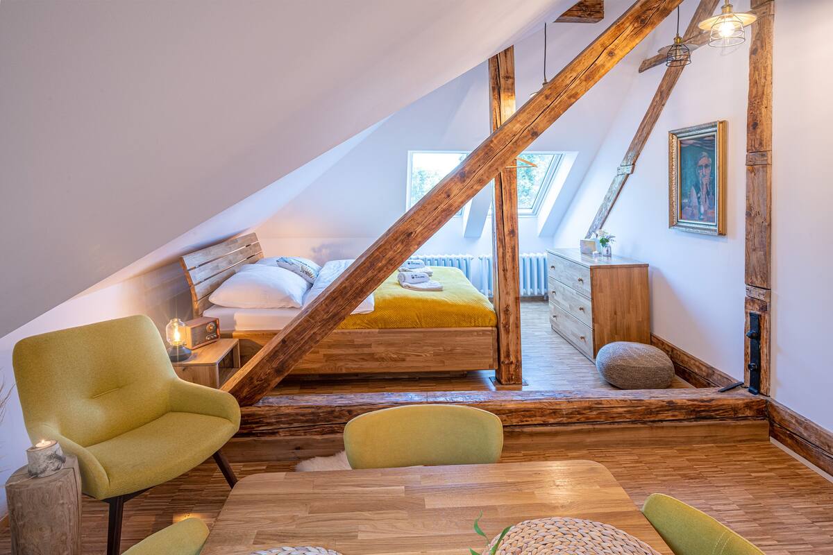 Une chambre à coucher aménagée dans un grenier, avec un plafond en pente, des poutres en bois, un lit doté d’un matelas jaune et un coin salon avec des fauteuils.