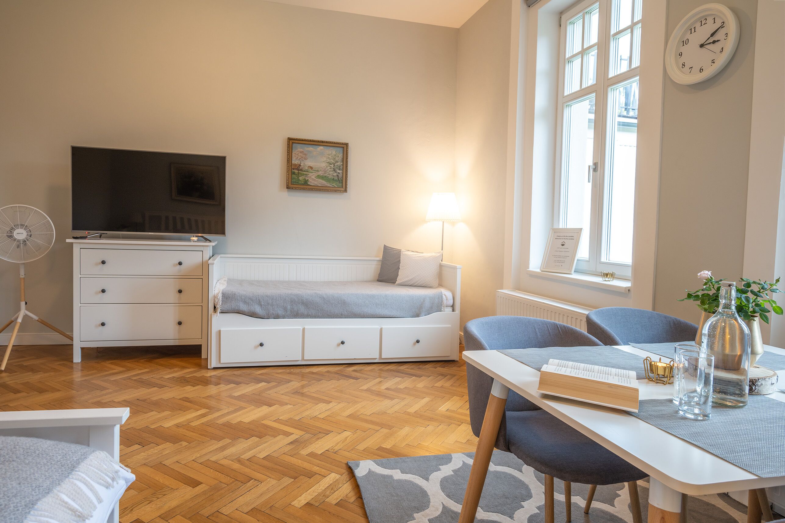 A modern room with a bed, a desk with a lamp, a TV, and a clock on the wall.
