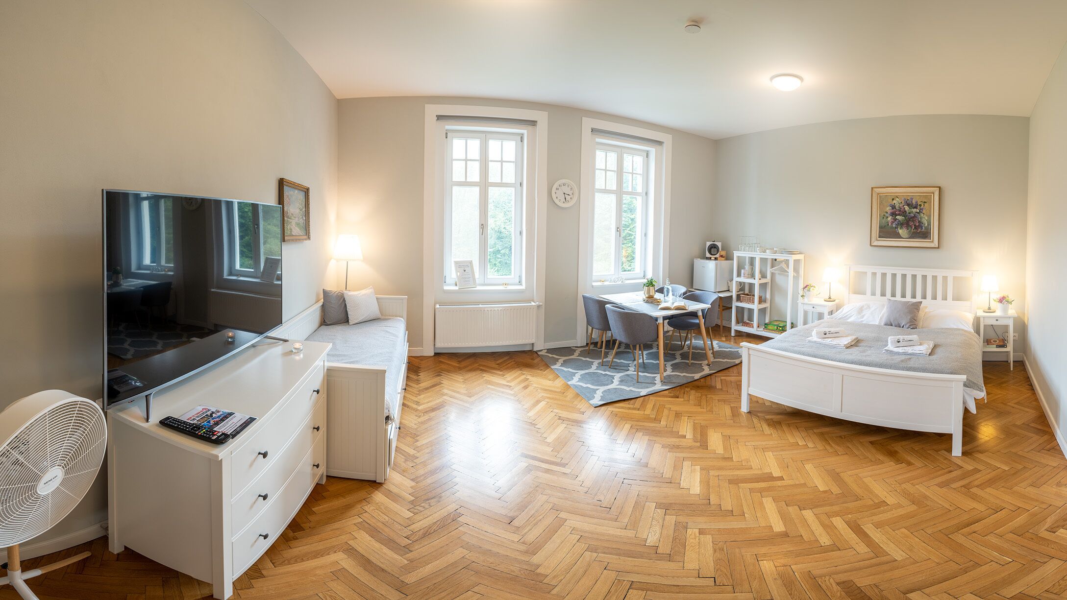 Quarto moderno e bem iluminado com televisão, escrivaninha, cama e área de refeições.