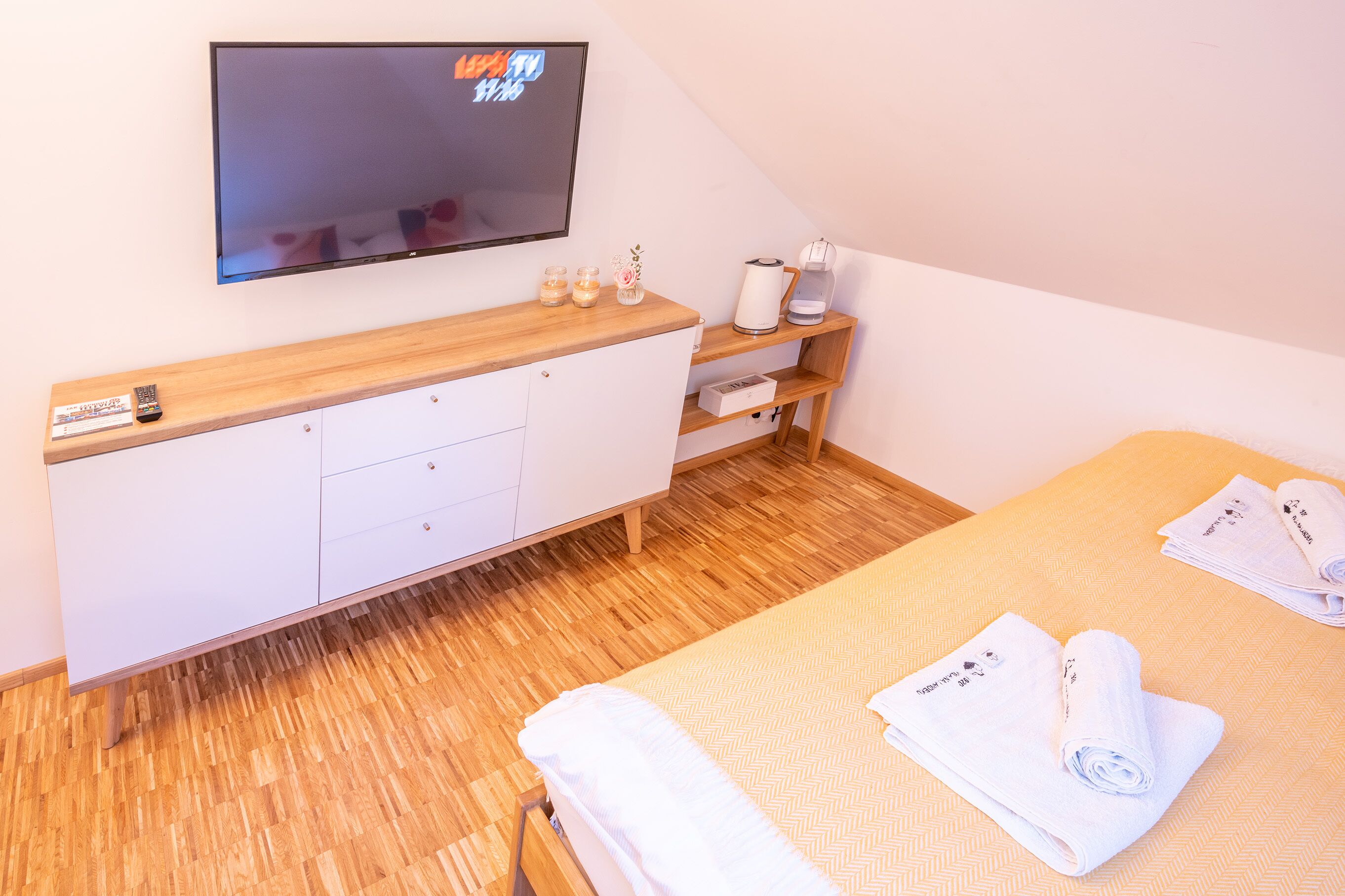Quarto de hotel com piso de madeira, cama com roupa branca, televisão fixada na parede, penteadeira de madeira e prateleira pequena com chaleira e chávenas.