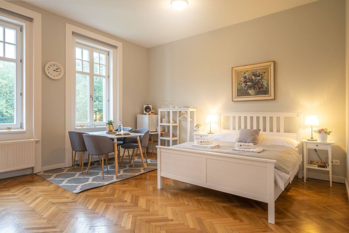 Une chambre à coucher comprenant un lit, des tables de chevet, un bureau avec des chaises, un radiateur et une fenêtre donnant sur un espace verdoyant.