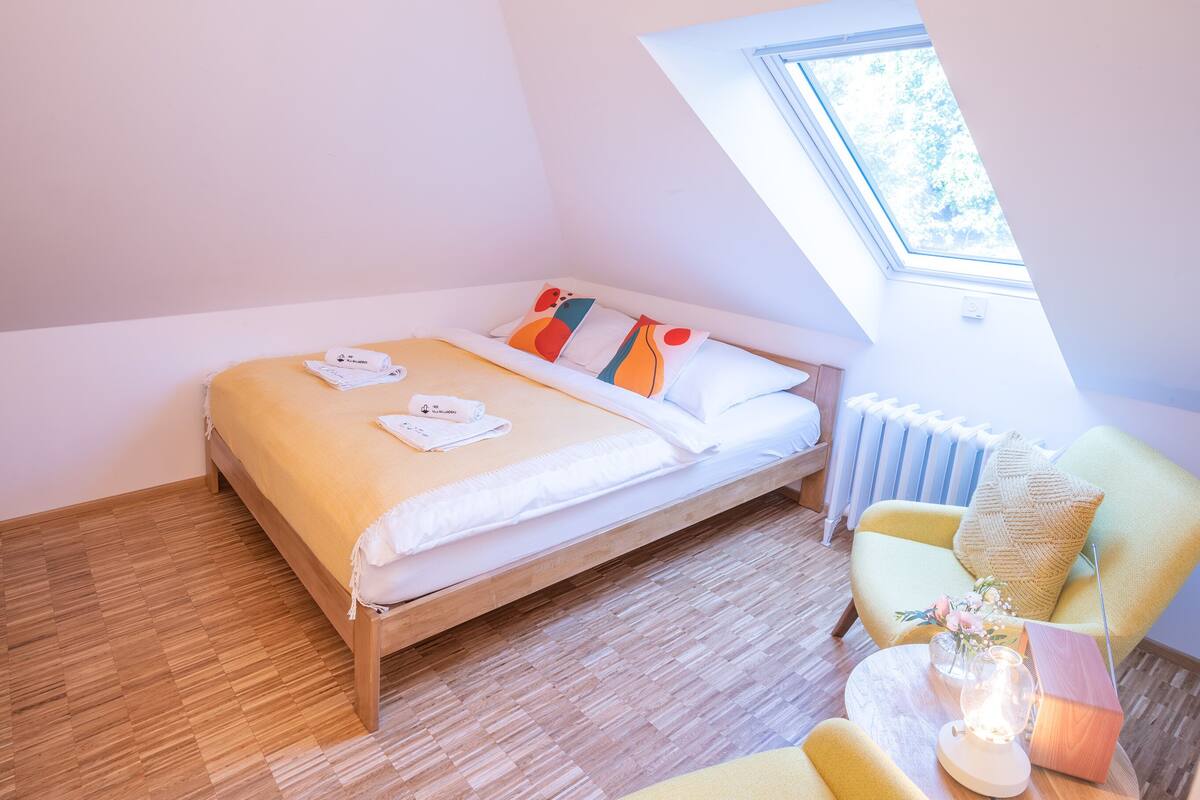Une chambre à coucher avec un lit en bois, un fauteuil jaune, une petite table avec une lampe et une lucarne.