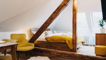A cozy attic bedroom with a sloped ceiling, wooden beams, a bed with a yellow blanket, a yellow armchair, and a white fur rug.