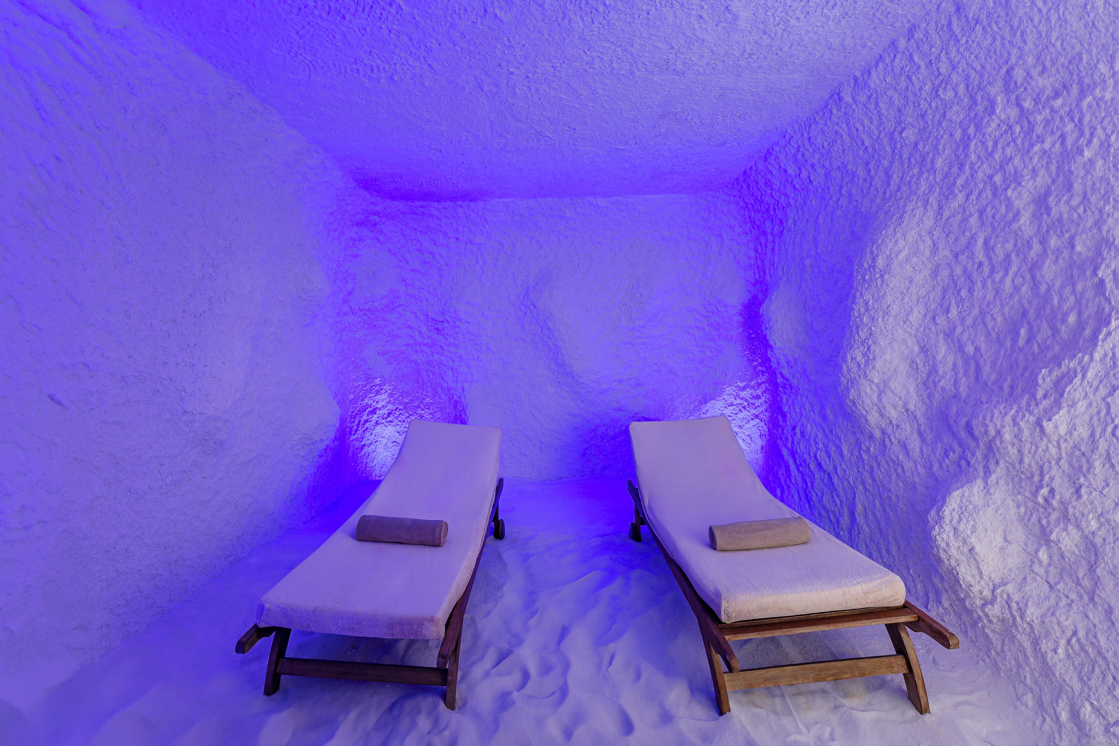 Two wooden lounge chairs with cushions in a room with textured walls and a blue light.