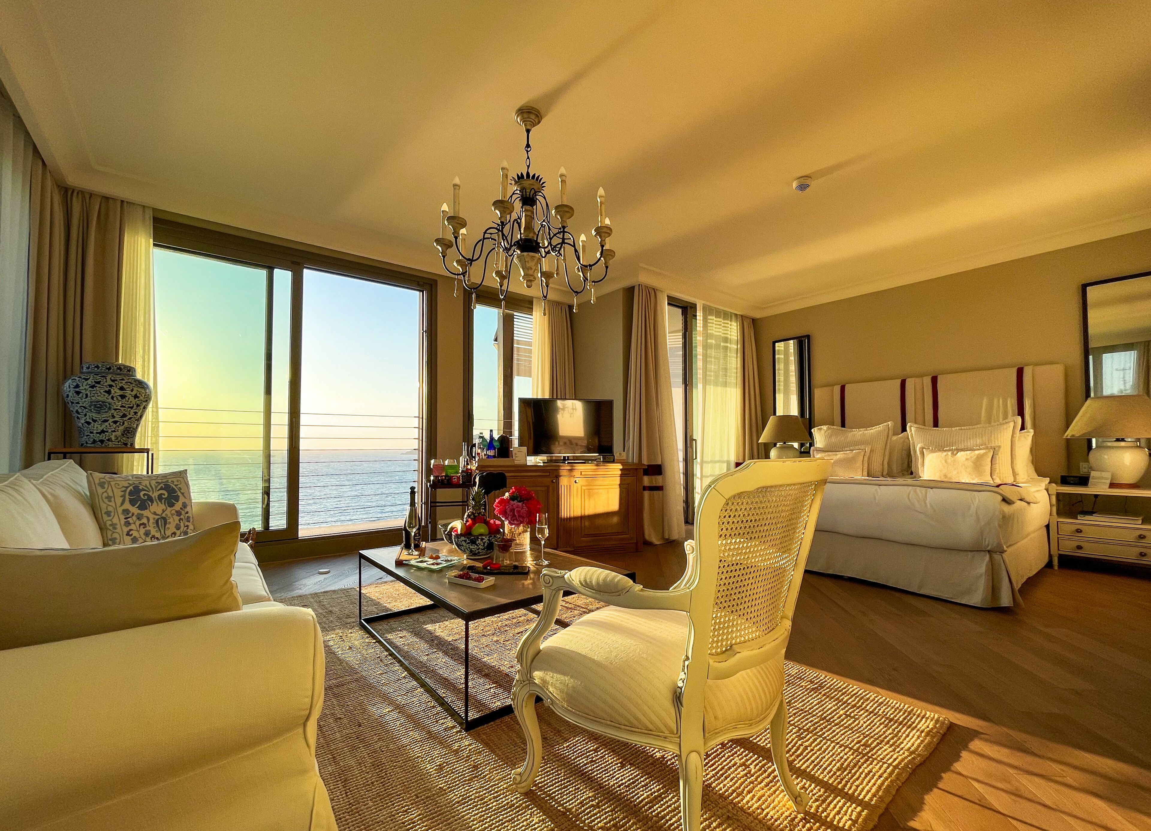 A hotel room with a large window overlooking the sea, a bed with white bedding, a sitting area with a sofa and armchair, a coffee table with flowers, and a chandelier.