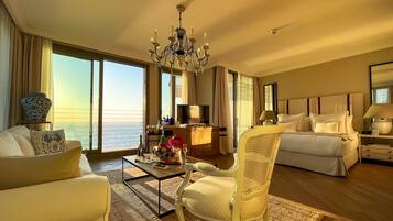 A hotel room with a large window overlooking the sea, a bed with white bedding, a sitting area with a sofa and armchair, a coffee table with flowers, and a chandelier.