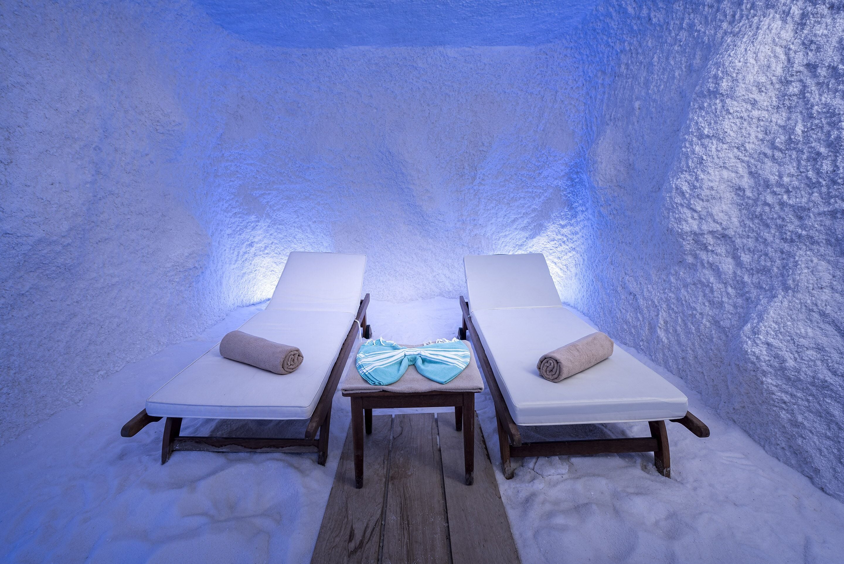 Two lounge chairs with towels, a wooden table, and a textured wall in a room with a blue hue.