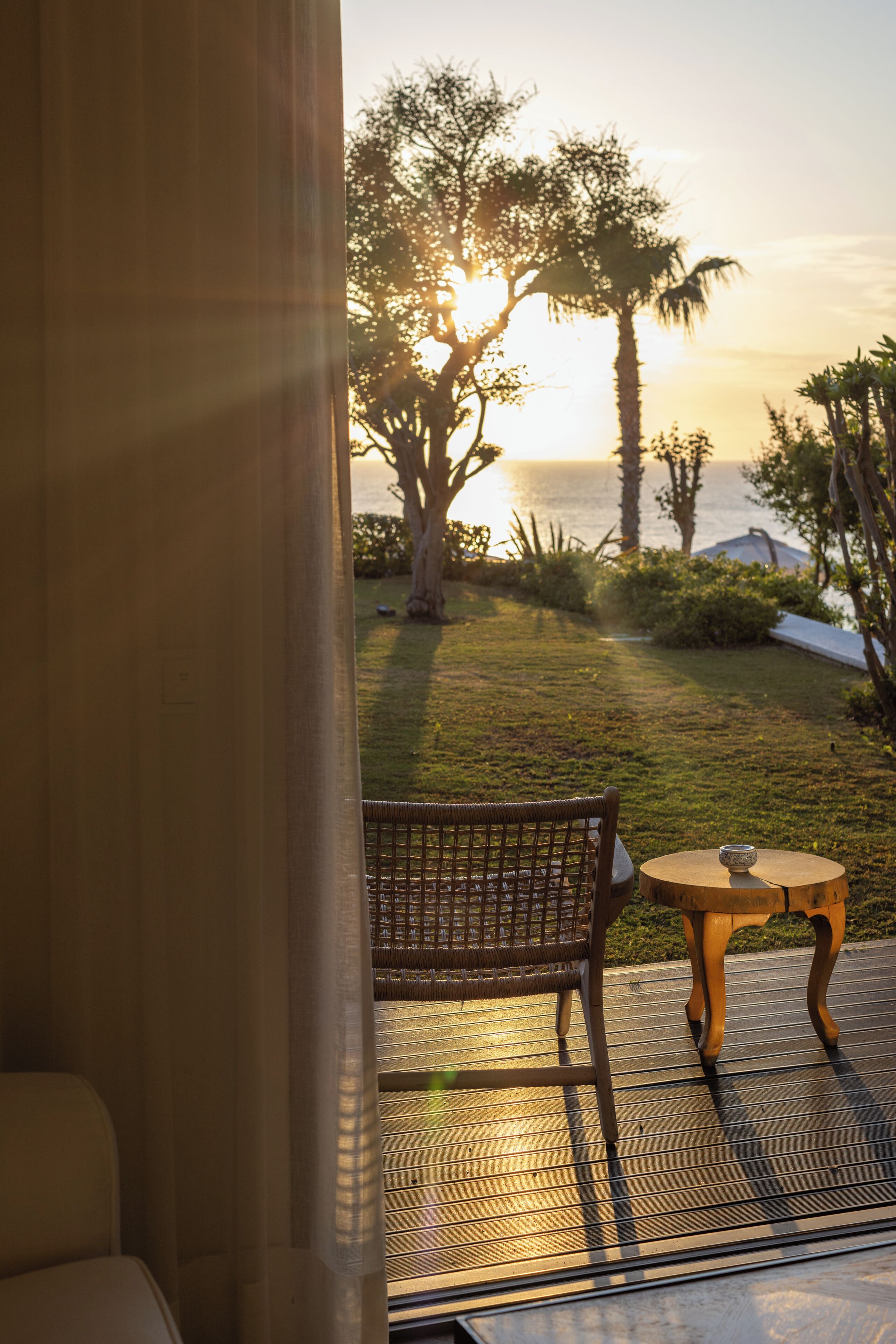 A room with a view of the sunset over the sea, a wicker chair, and a small round table with a cup.