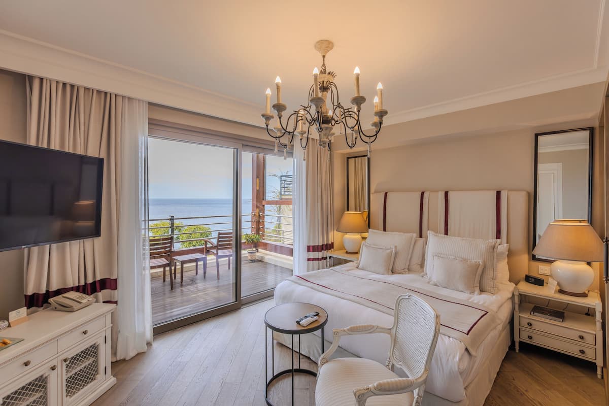 A hotel room with a large bed, a television, a chandelier, a balcony with a view of the sea, and a dining area with a table and chairs.
