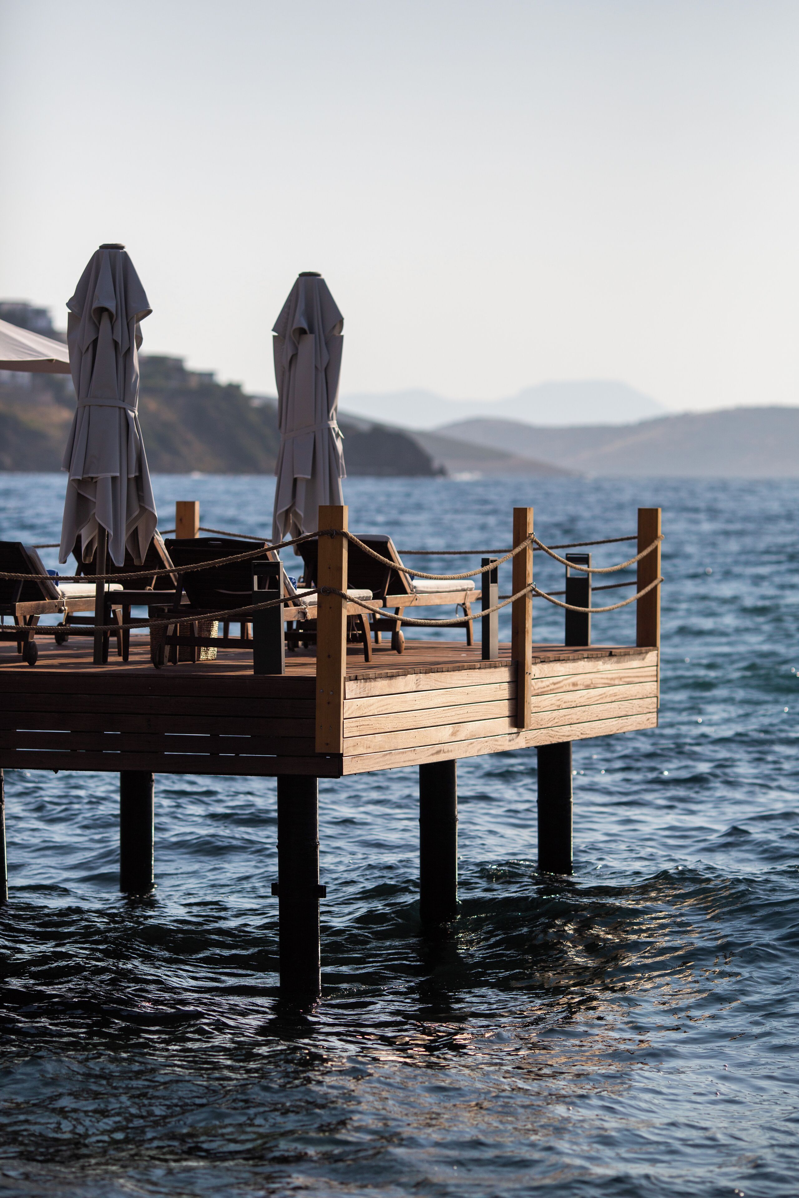 A wooden pier with lounge chairs and umbrellas, extending over calm water with mountains in the background.