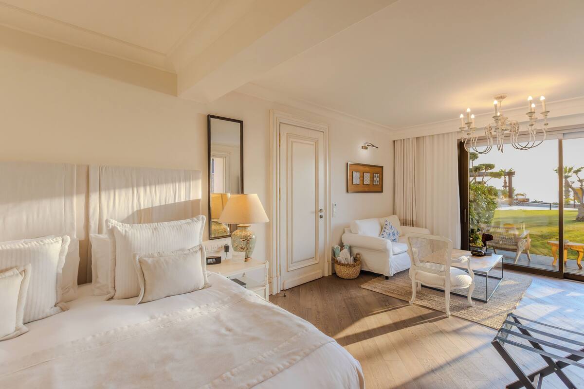 A hotel room with a large bed, a sitting area with a chair and table, a door leading to a balcony, and a chandelier.