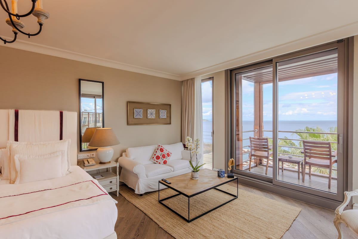 A hotel room with a bed, a sofa, a coffee table, a lamp, a vase with flowers, a nightstand, a mirror, and a balcony with a view of the ocean.