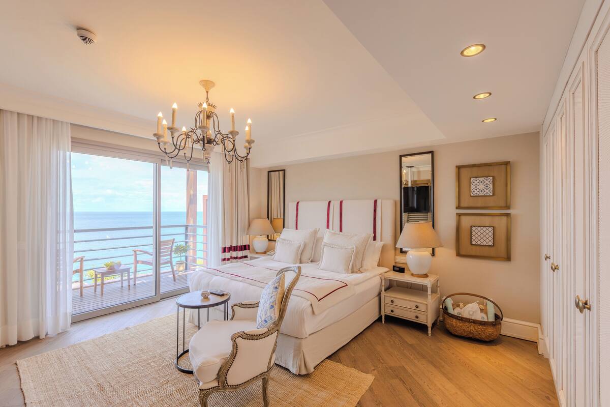 A spacious bedroom with a large bed, a sitting area with a chair, a small table, and a view of the ocean through a sliding door.