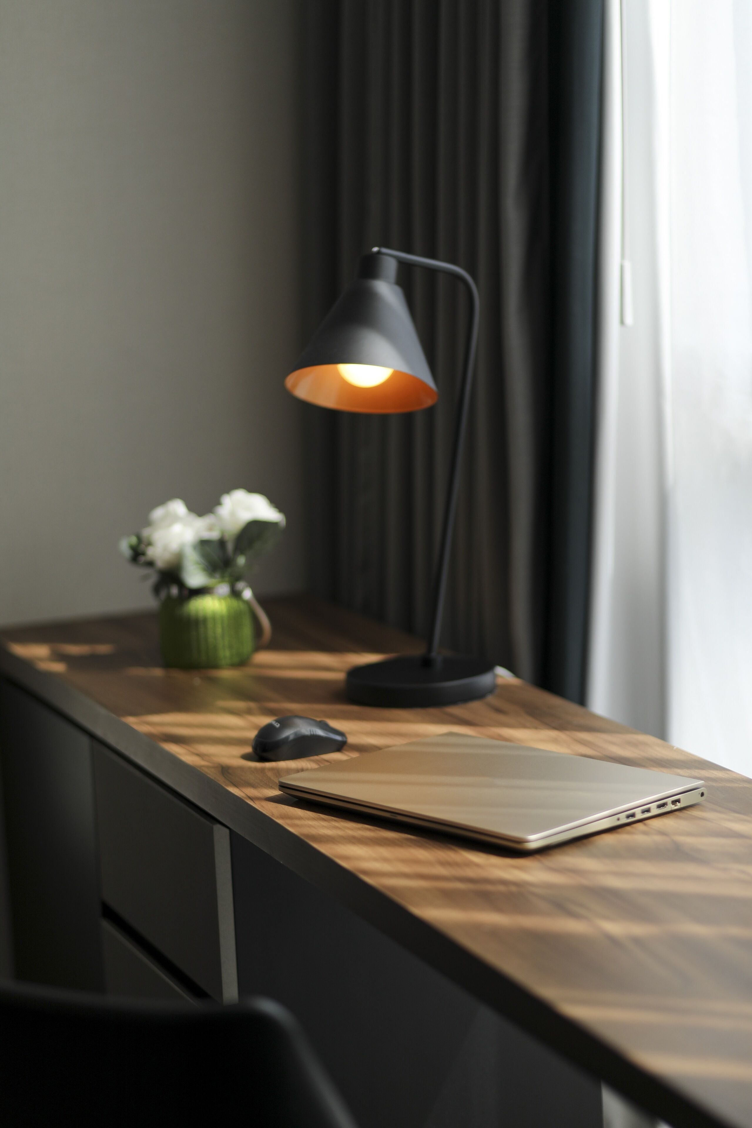 Een houten bureau met een laptop, een zwarte bureaulamp en een groene vaas met witte bloemen.