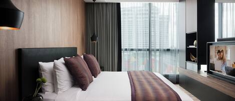 Habitación de hotel moderna con una cama grande, una mesita de noche con un jarrón y una vela, un ventanal con vistas a la ciudad y un televisor a la derecha.