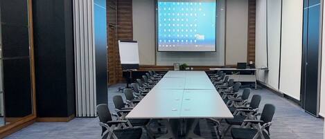 Un salón de conferencias con una larga mesa, sillas, una pantalla de proyector y una pizarra.